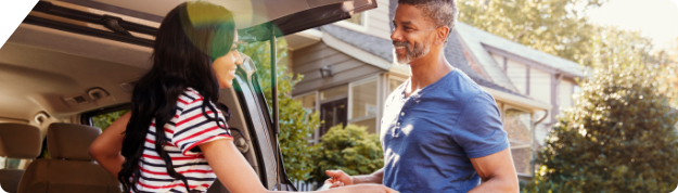 A couple smile at each other as they unload the trunk of their vehicle parked outside their home. 